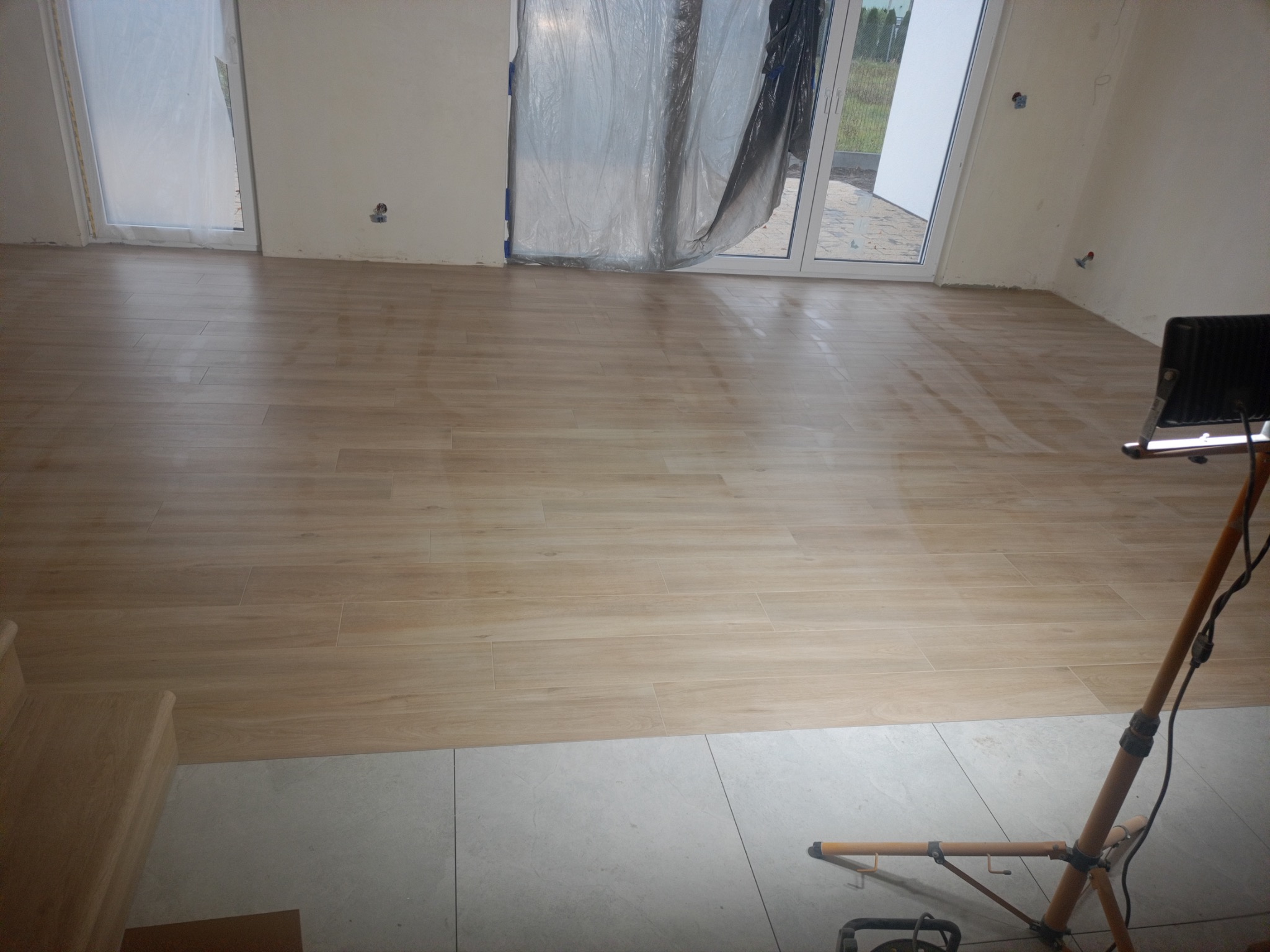 Jasne panele podłogowe ułożone w nowym pomieszczeniu, widoczna lampa budowlana i zabezpieczone folią okna.