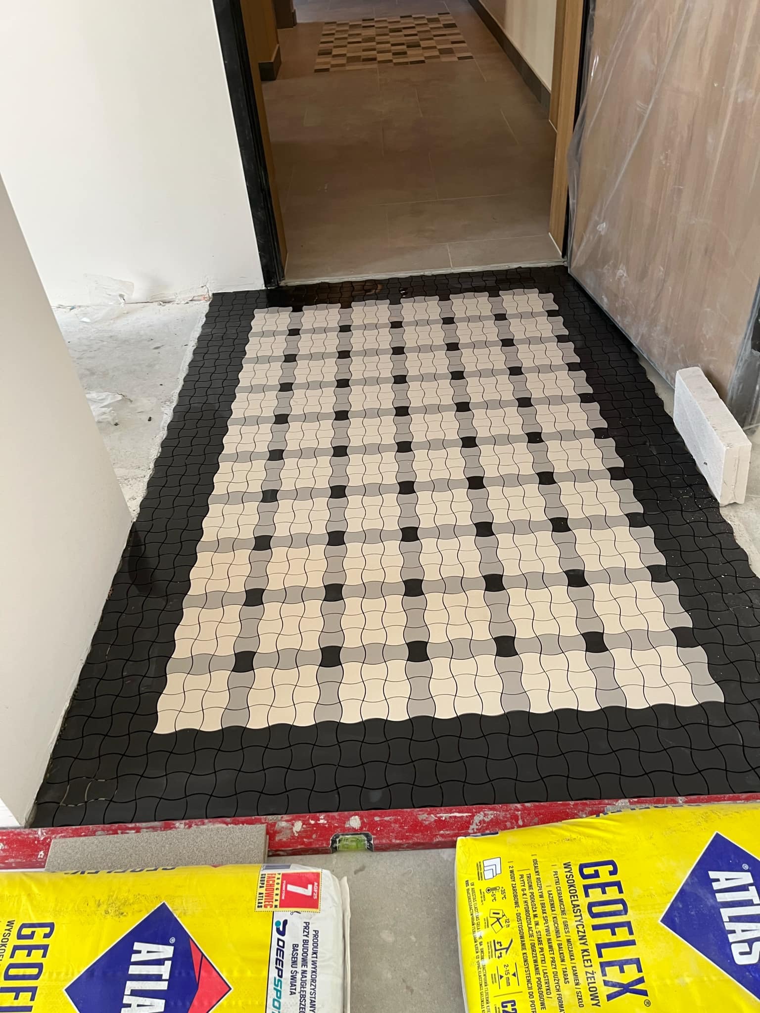 Układanie mozaiki podłogowej w odcieniach szarości i beżu, z czarną obwódką, widoczne worki kleju Atlas Geoflex i poziomica, w tle fragment ułożonej podłogi w innym pomieszczeniu.
