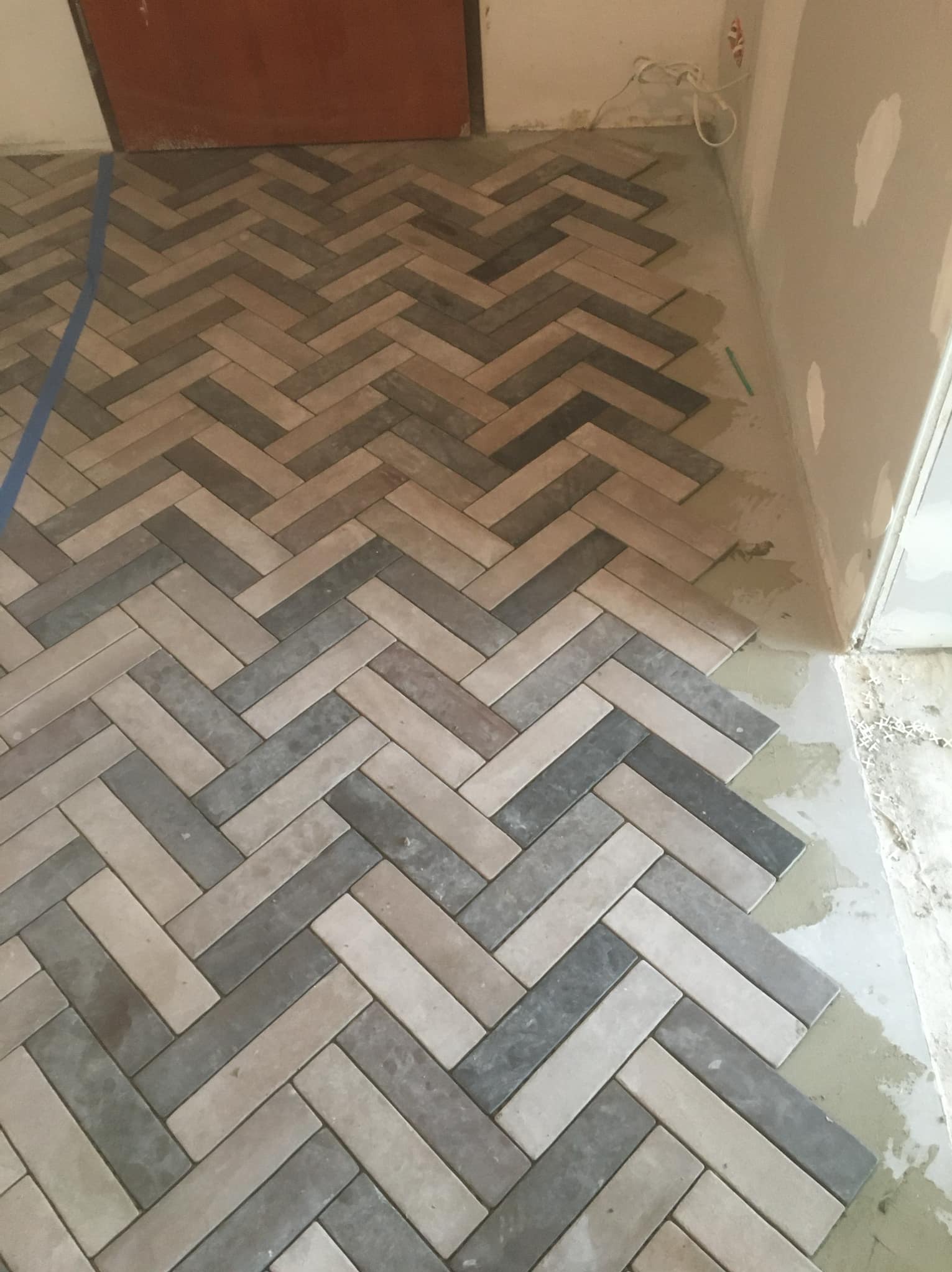 Układanie podłogi z płytek w jodełkę, widoczne świeże fugi i taśma malarska zabezpieczająca ścianę.