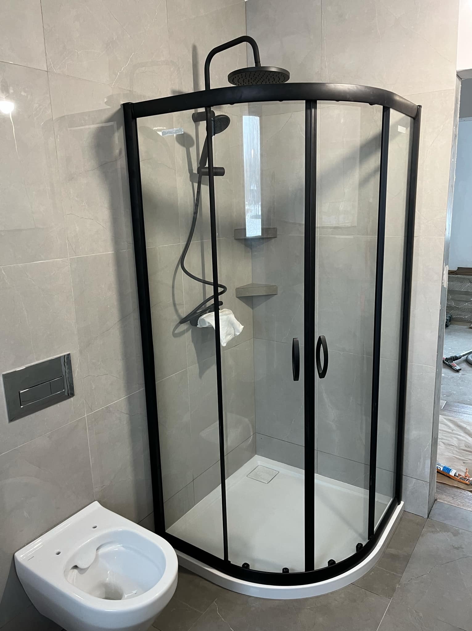 Nowoczesna kabina prysznicowa z czarnymi profilami i deszczownicą, zamontowana w łazience wykończonej szarymi płytkami imitującymi marmur. Umywalka podwieszana widoczna w lewym dolnym rogu.