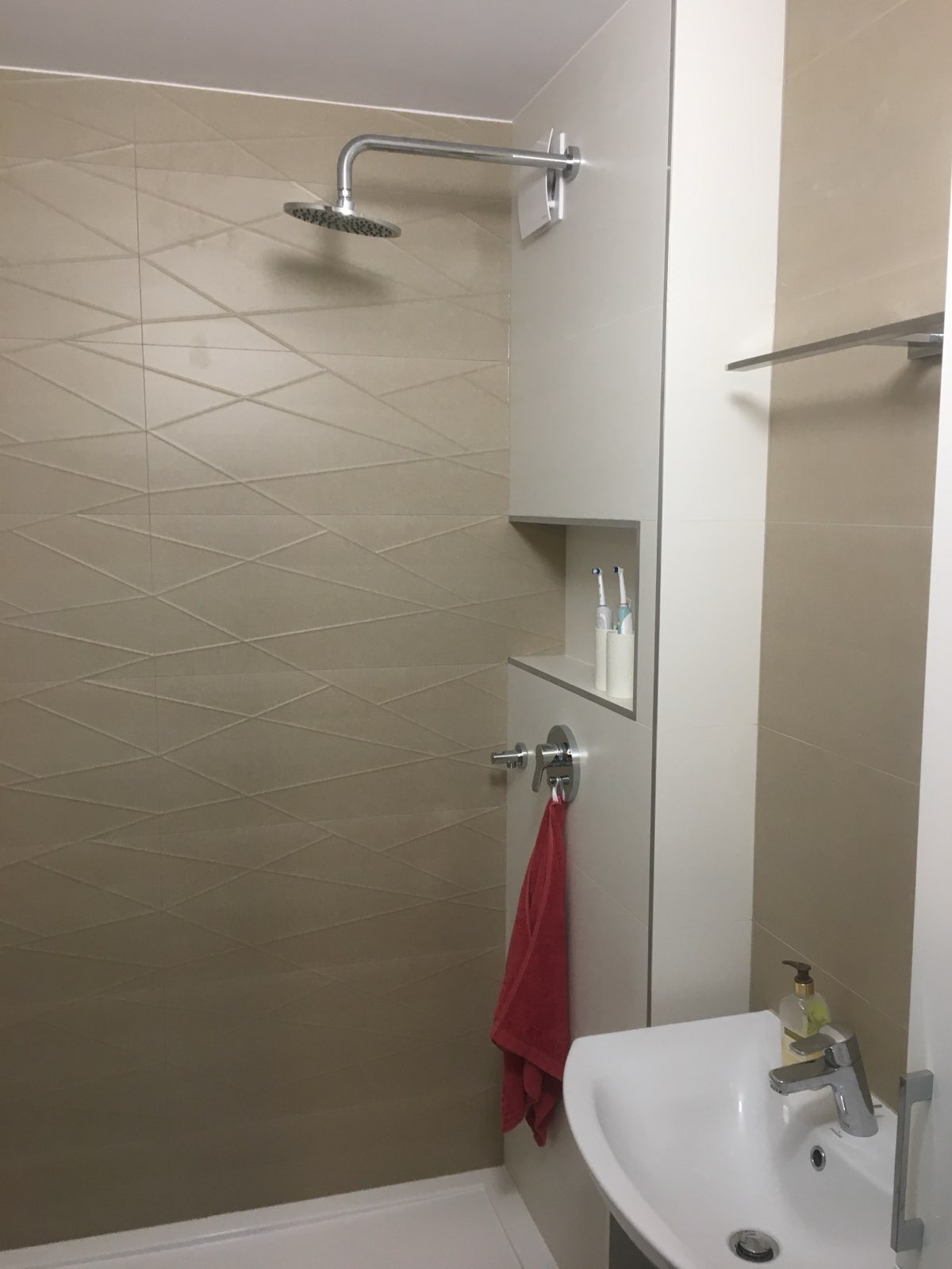 Nowoczesna kabina prysznicowa z beżowymi płytkami o geometrycznym wzorze, chromowaną armaturą i wnęką z elektrycznymi szczoteczkami do zębów, uzupełniona o czerwoną wiszącą szmatkę i umywalkę...