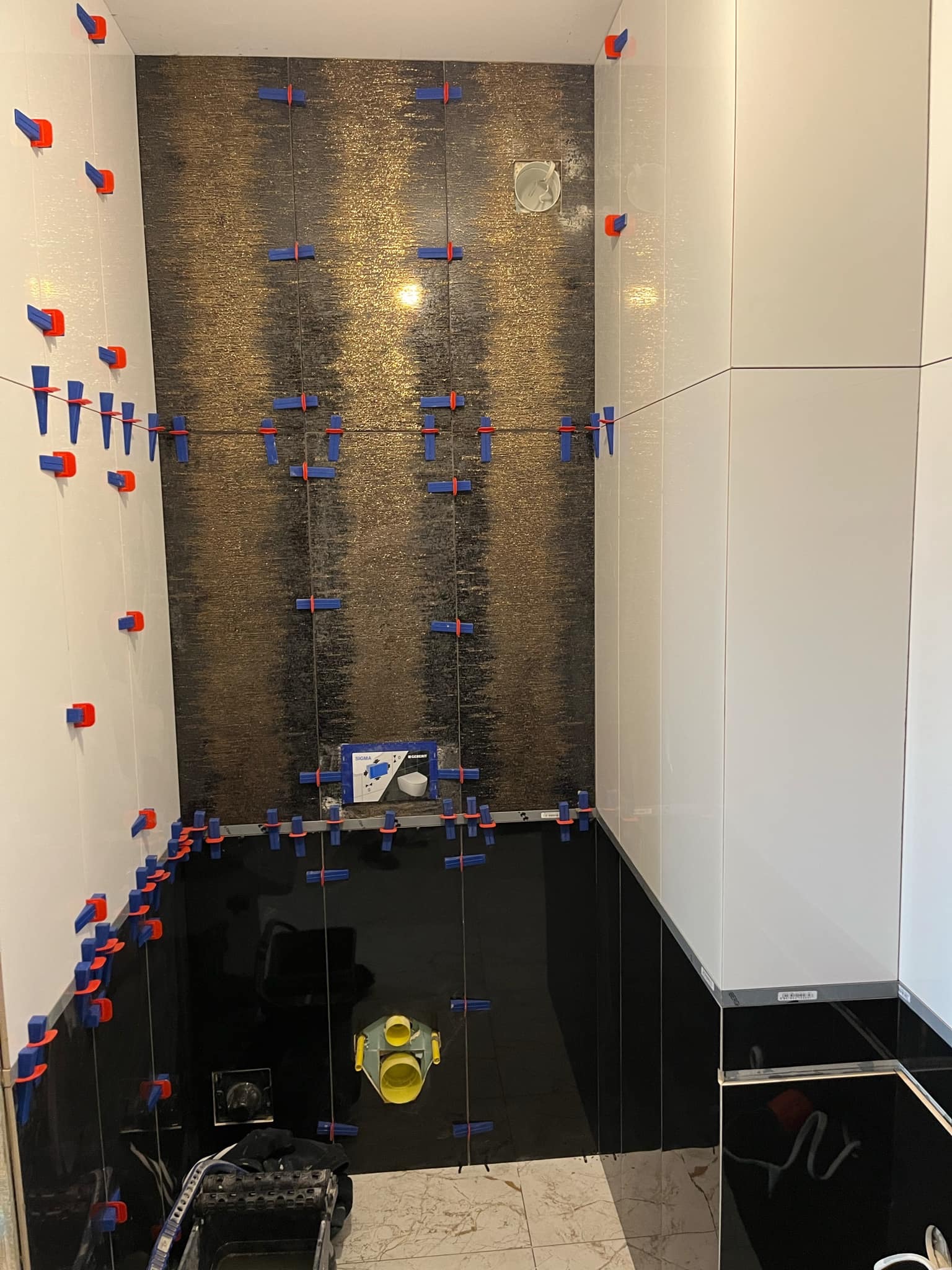 Ściany łazienki w trakcie układania płytek, widoczne narzędzia i system poziomowania płytek, czarne i złote płytki na ścianie, białe płytki po bokach.