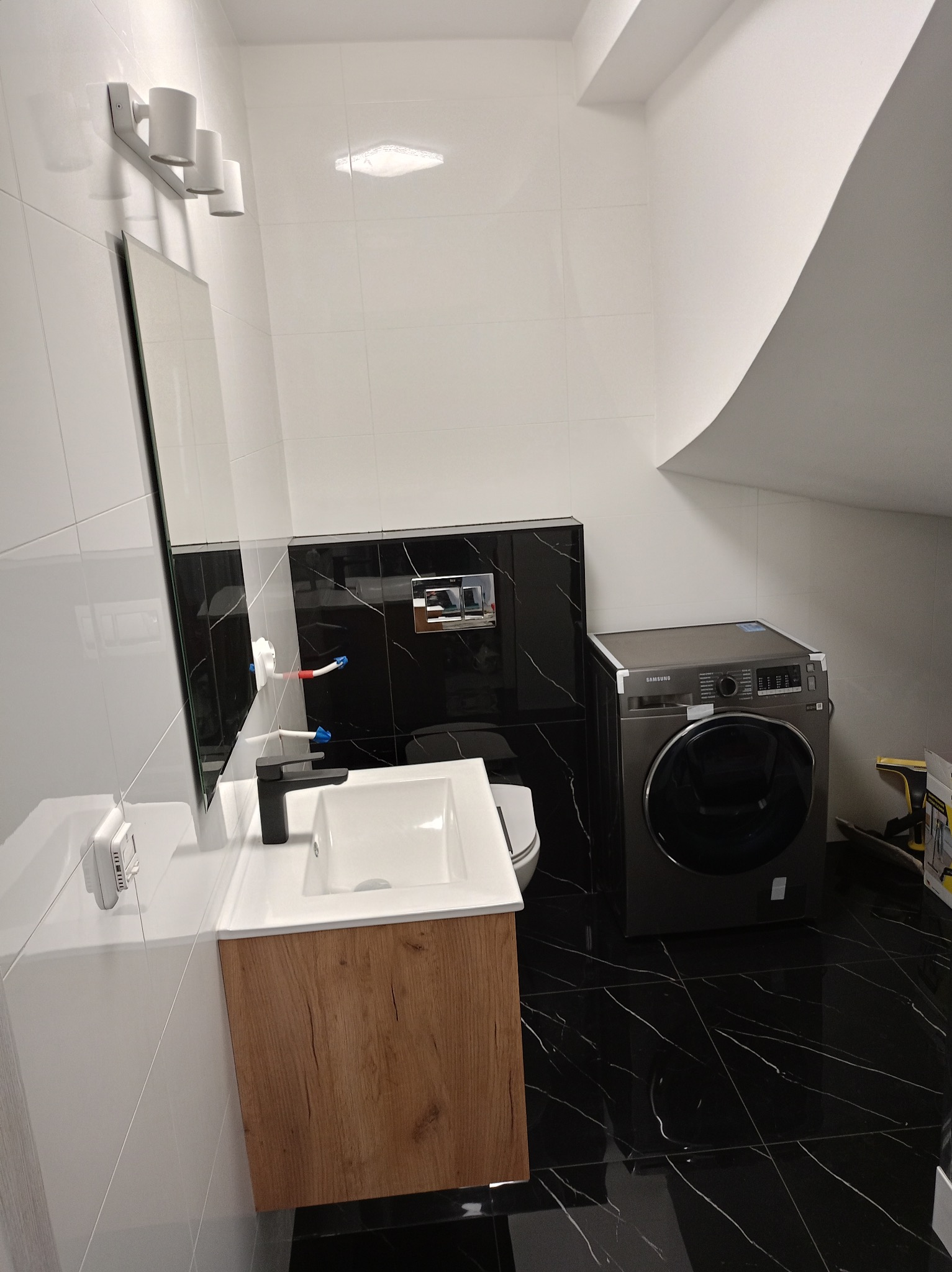 Wykończona łazienka w stylu nowoczesnym z czarno-białymi płytkami imitującymi marmur, umywalką z szafką w kolorze drewna, pralką Samsung i oświetleniem punktowym nad lustrem.