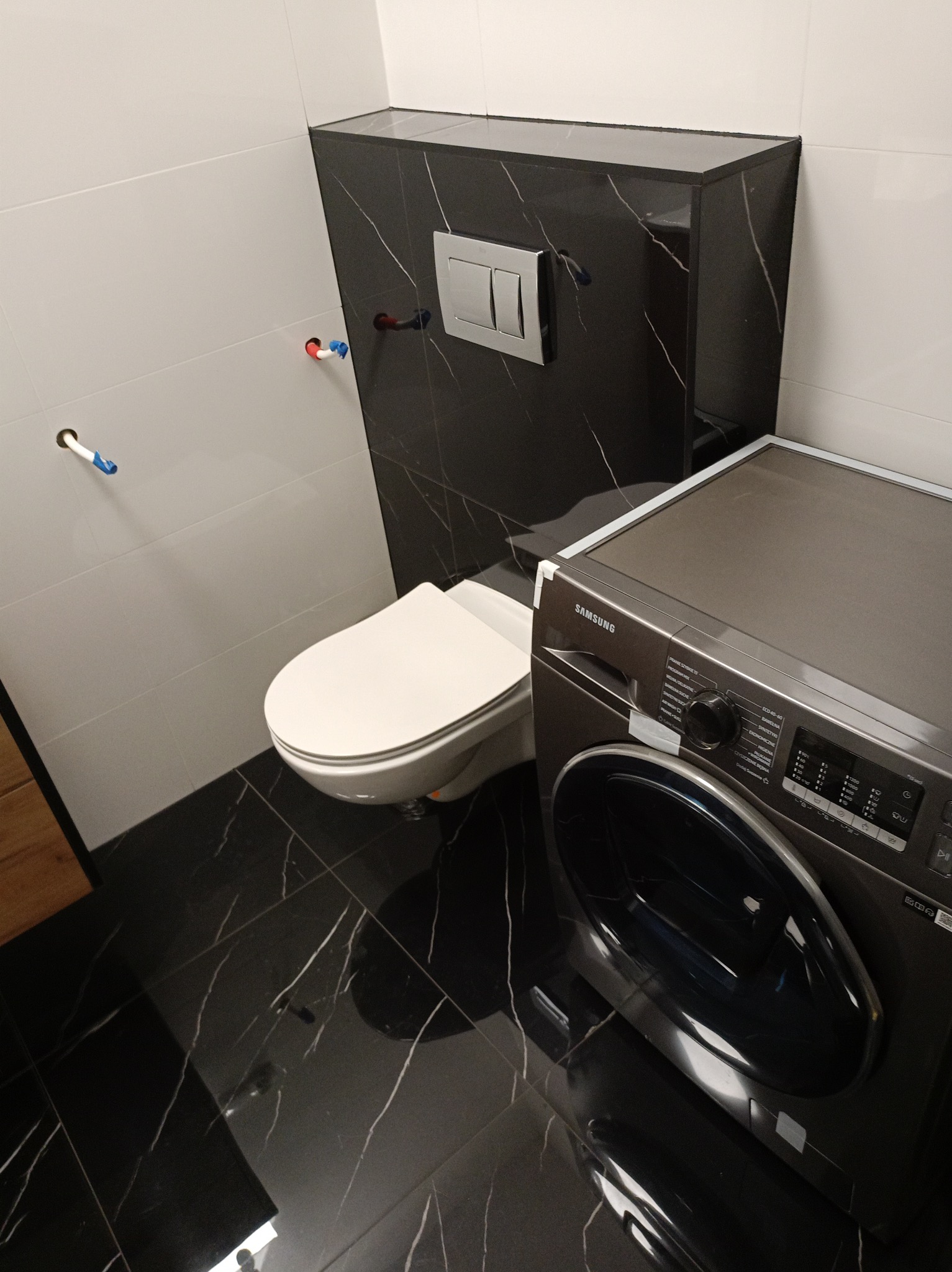 Wykończona łazienka z czarno-białymi płytkami, wiszącą toaletą i pralką Samsung w kolorze grafitowym.