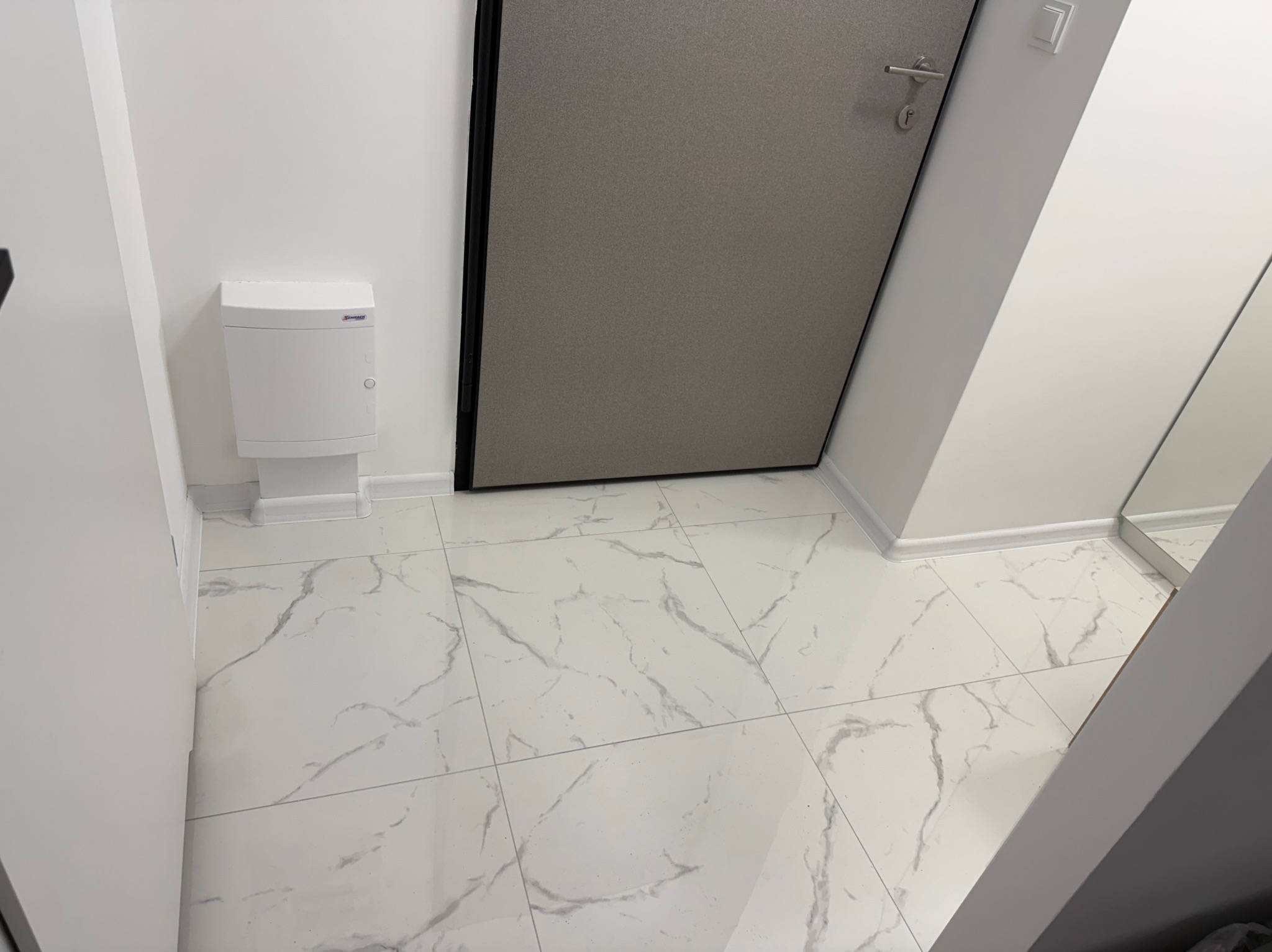 Białe, marmurkowe płytki podłogowe w korytarzu, z widoczną szafką na ścianie i drzwiami wejściowymi.