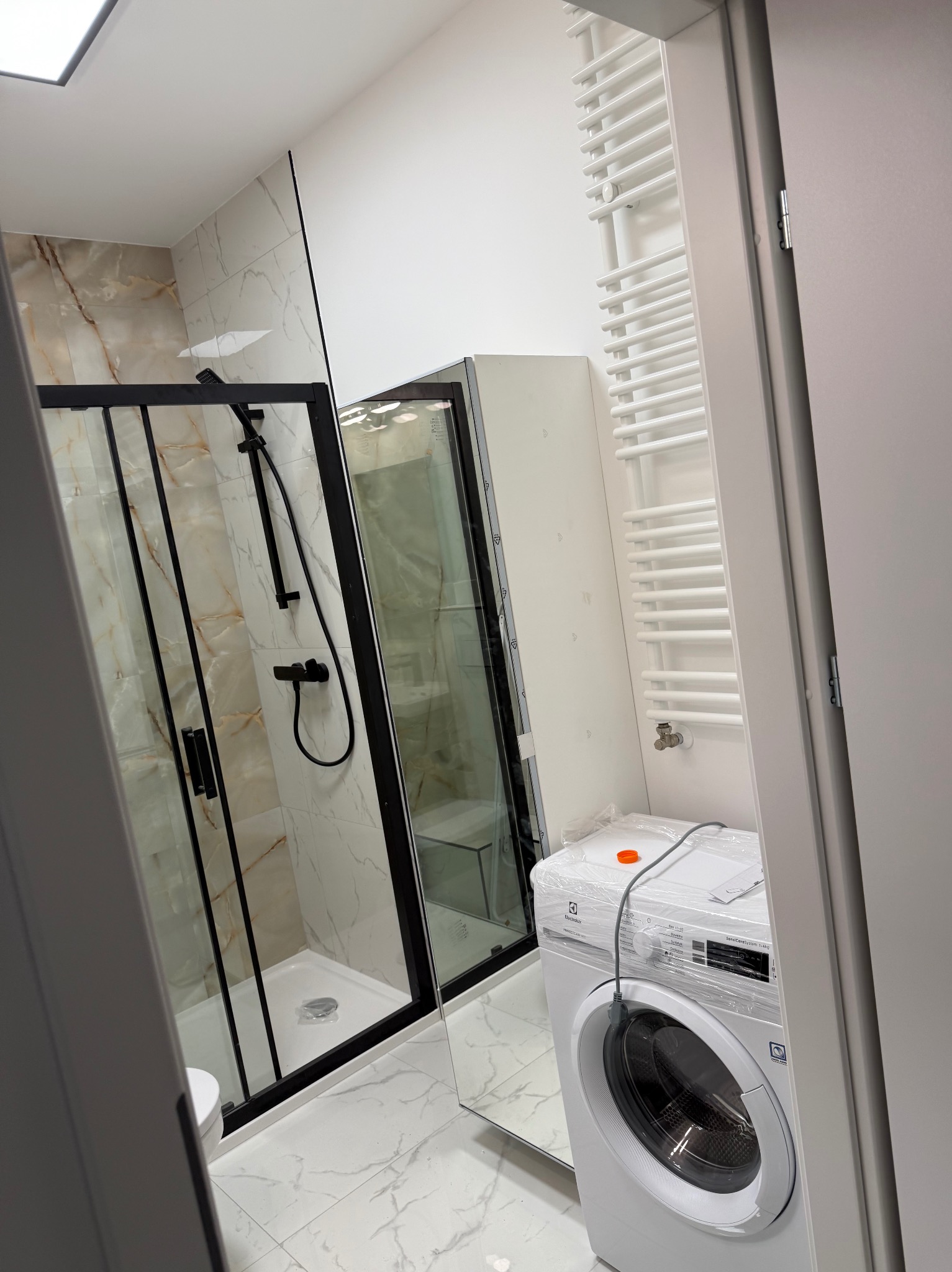 Nowoczesna łazienka z kabiną prysznicową w czarnej ramie, marmurowe płytki na ścianach, biała pralka Electrolux jeszcze w folii ochronnej i grzejnik drabinkowy.