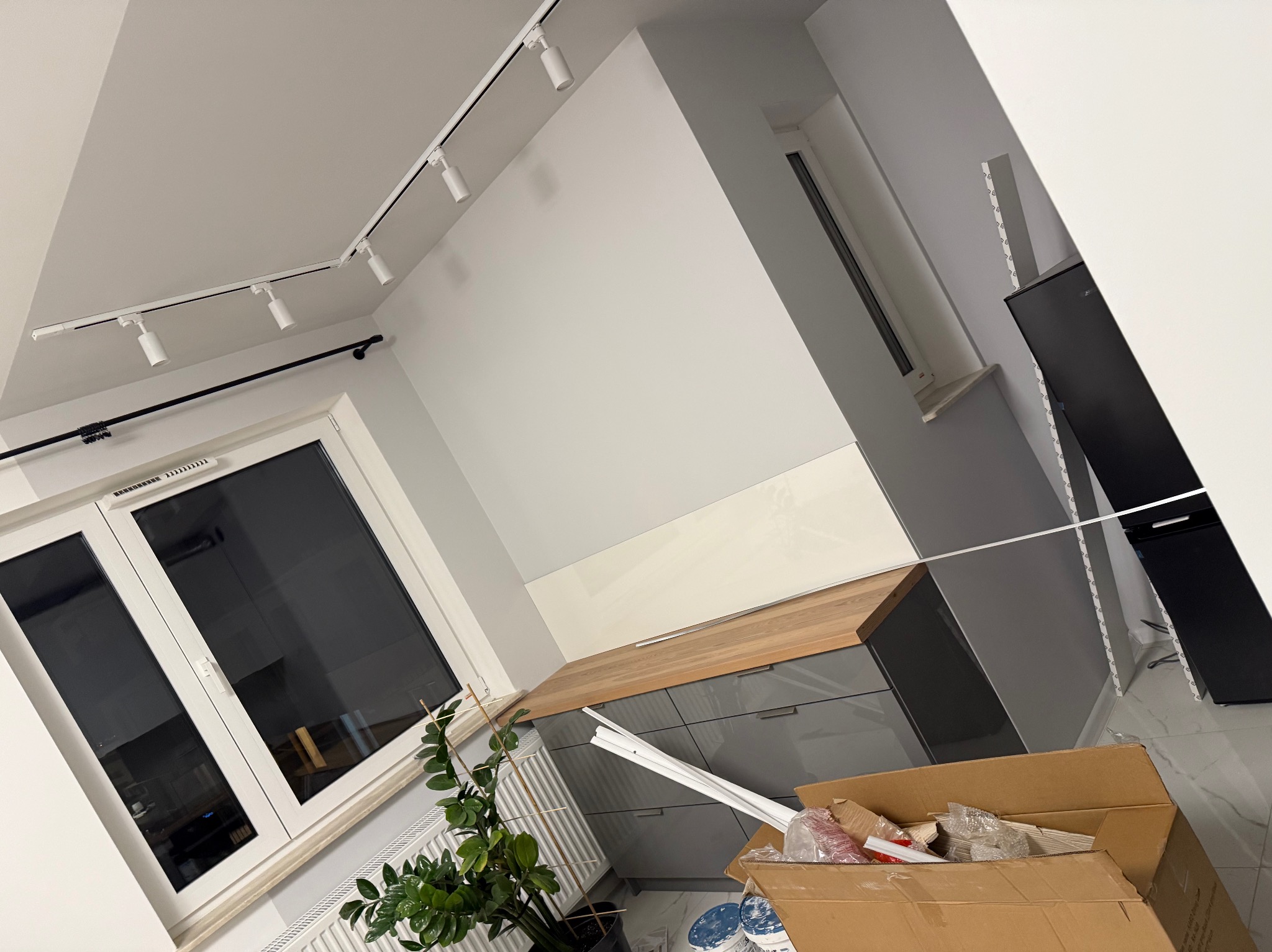 Montaż szarych szafek kuchennych z drewnianym blatem, widoczne listwy przypodłogowe i karton z materiałami montażowymi, oświetlenie szynowe na suficie.