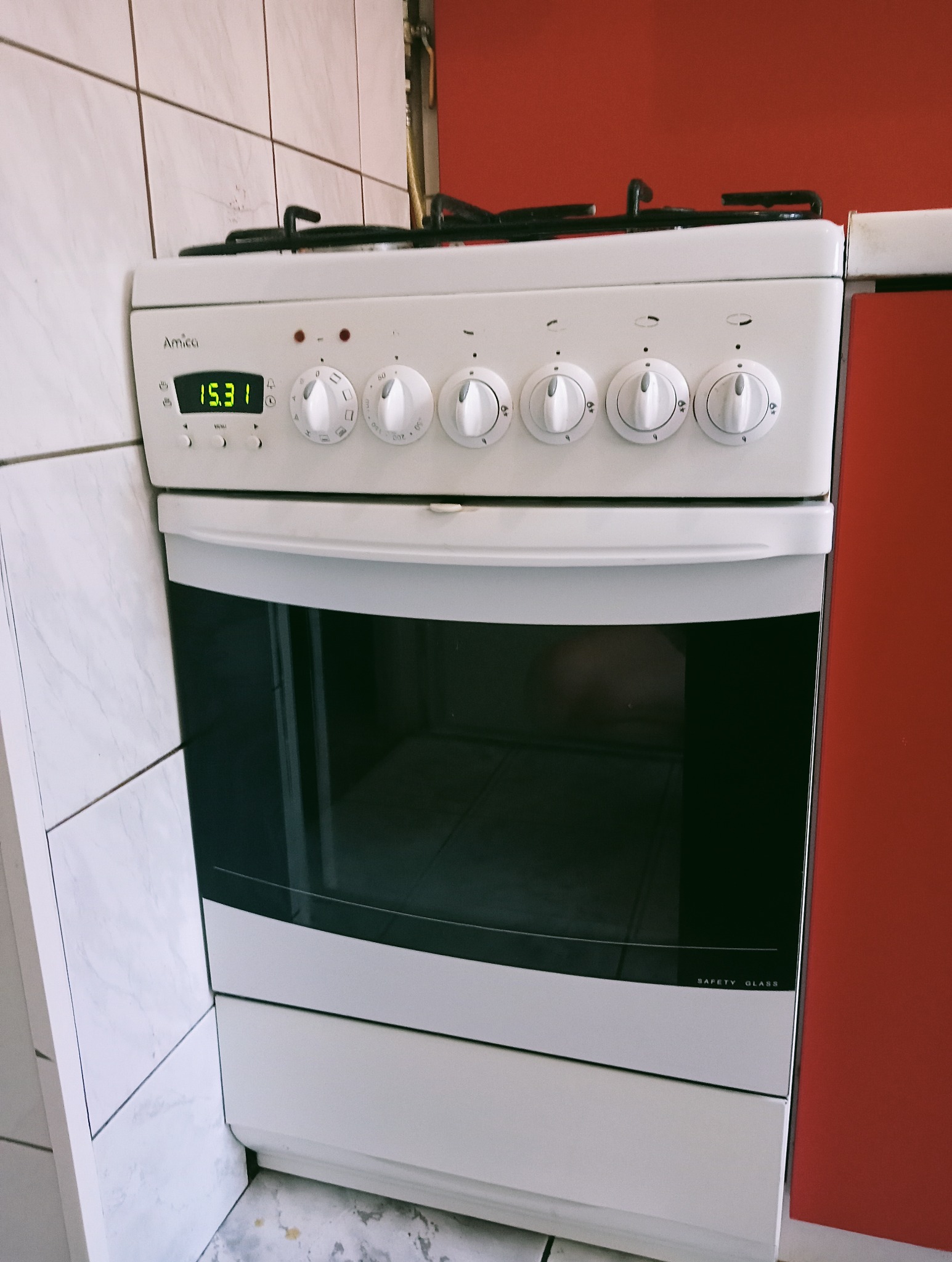 Biała kuchenka gazowa marki Amica z wyświetlaczem wskazującym godzinę 15:31, umieszczona między białą ścianą wyłożoną płytkami a czerwoną ścianą