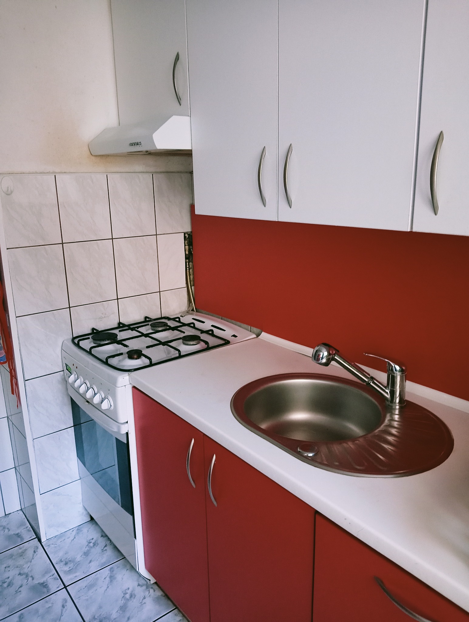 Kuchnia z czerwonymi szafkami, białym blatem i zlewozmywakiem ze stali nierdzewnej, widok na kuchenkę gazową i okap.