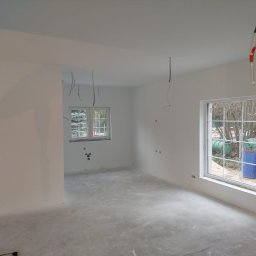 MShouse - Wnętrze pokoju w trakcie przygotowania do prac wykończeniowych: świeżo wyszpachlowane ściany, surowy beton na podłodze, wystające przewody elektryczne i instalacja wodna w tle okna.