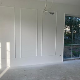 MShouse - Białe ściany z dekoracyjnymi listwami w nowym pokoju, widoczne wystające kable elektryczne i duże okno z widokiem na zieleń, podłoga w stanie surowym.