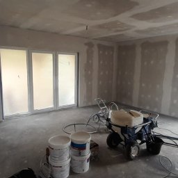 MShouse - Pokój w trakcie szpachlowania maszynowego, widoczne ściany z nałożoną pierwszą warstwą gładzi, agregat malarski, drabina i wiaderka z masą szpachlową.