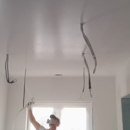 MShouse - Malowanie natryskowe sufitu w pomieszczeniu z widocznymi przewodami elektrycznymi i otworem na oświetlenie, pracownik w masce ochronnej i kombinezonie maluje sufit na biało.