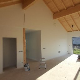 MShouse - Wnętrze w trakcie prac wykończeniowych: pomalowane ściany, drewniana konstrukcja dachu, surowe otwory drzwiowe, drabina i wiadro z farbą na podłodze.