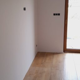 MShouse - Jasne, drewnopodobne płytki podłogowe ułożone w nowym pomieszczeniu z gładkimi, białymi ścianami, widoczne gniazdka elektryczne i klimatyzator, przez okno widać zieleń.