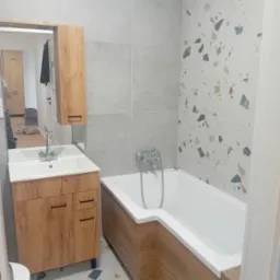 Wykończona łazienka w Bielawie z wanną obudowaną drewnopodobnymi panelami, umywalką z szafką w tym samym stylu, szarą glazurą i ścianą z wzorem terrazzo.