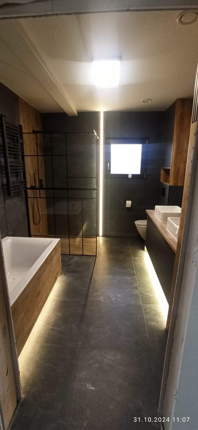 Nowoczesna łazienka z szarymi płytkami podłogowymi i ściennymi, drewnianymi akcentami, podświetleniem LED pod szafką umywalkową i pionowym paskiem świetlnym obok kabiny prysznicowej.