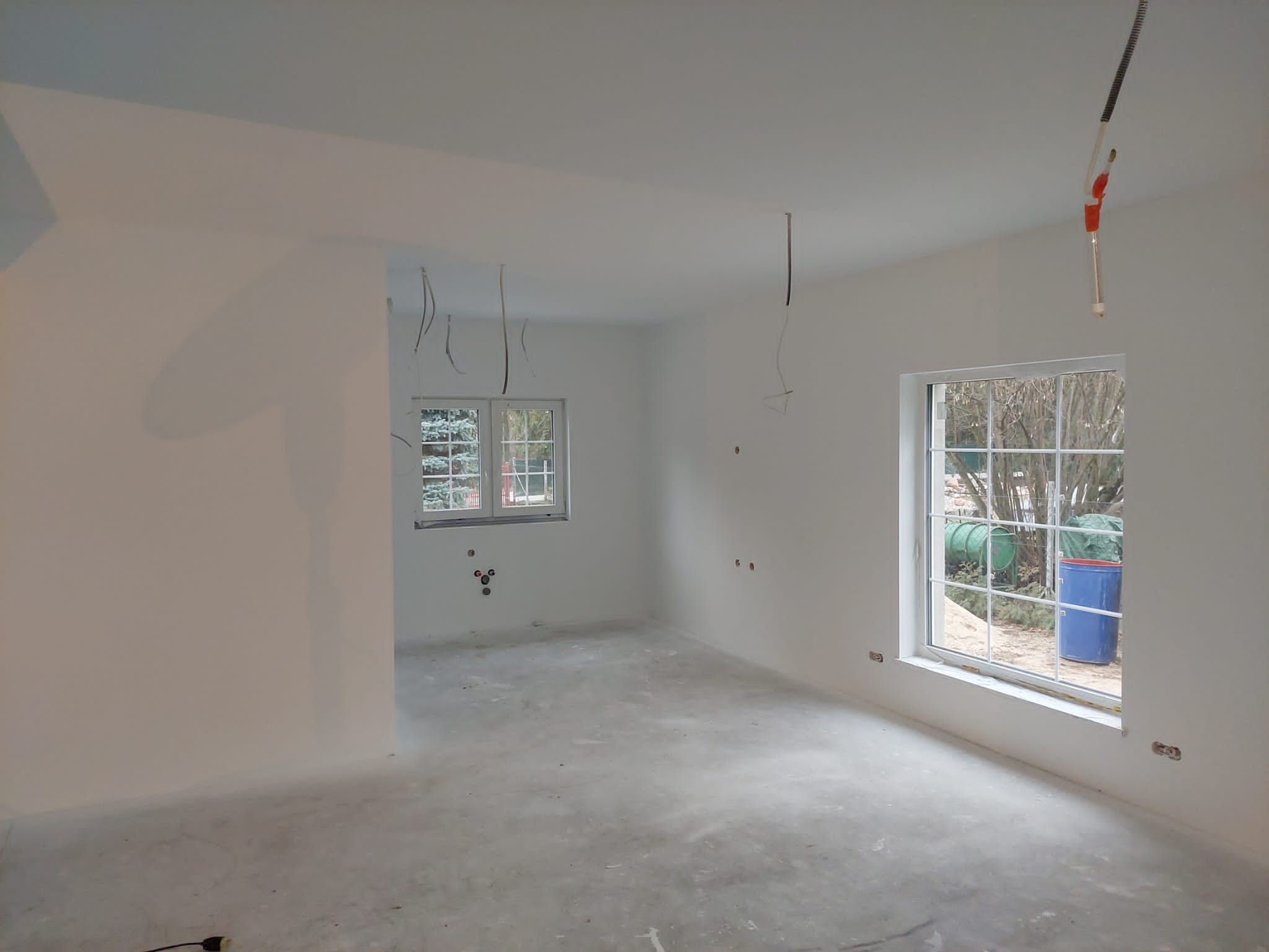 Wnętrze pokoju w trakcie przygotowania do prac wykończeniowych: świeżo wyszpachlowane ściany, surowy beton na podłodze, wystające przewody elektryczne i instalacja wodna w tle okna.
