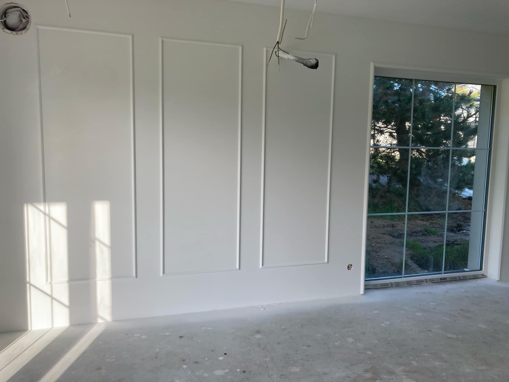 Białe ściany z dekoracyjnymi listwami w nowym pokoju, widoczne wystające kable elektryczne i duże okno z widokiem na zieleń, podłoga w stanie surowym.