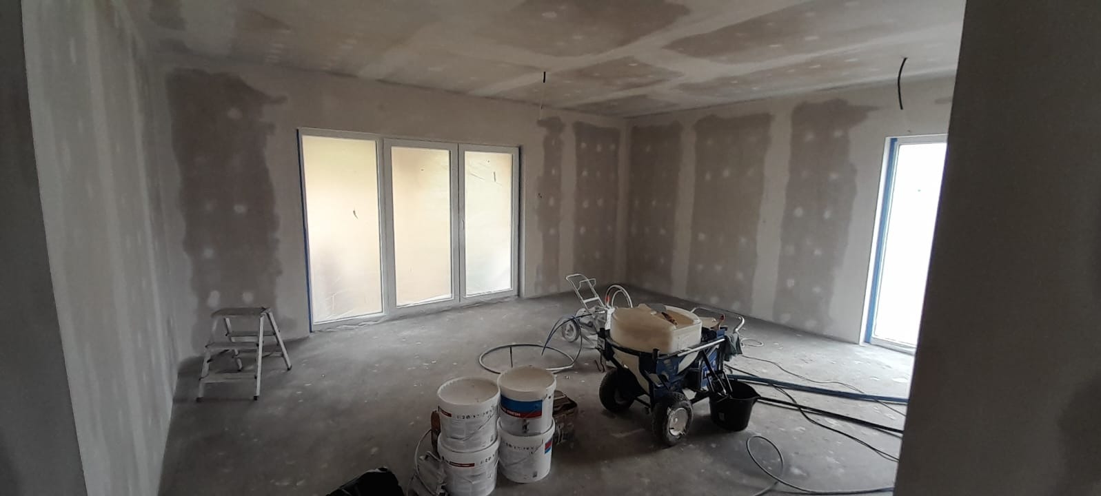 Pokój w trakcie szpachlowania maszynowego, widoczne ściany z nałożoną pierwszą warstwą gładzi, agregat malarski, drabina i wiaderka z masą szpachlową.