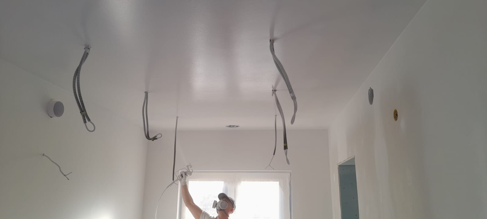 Malowanie natryskowe sufitu w pomieszczeniu z widocznymi przewodami elektrycznymi i otworem na oświetlenie, pracownik w masce ochronnej i kombinezonie maluje sufit na biało.