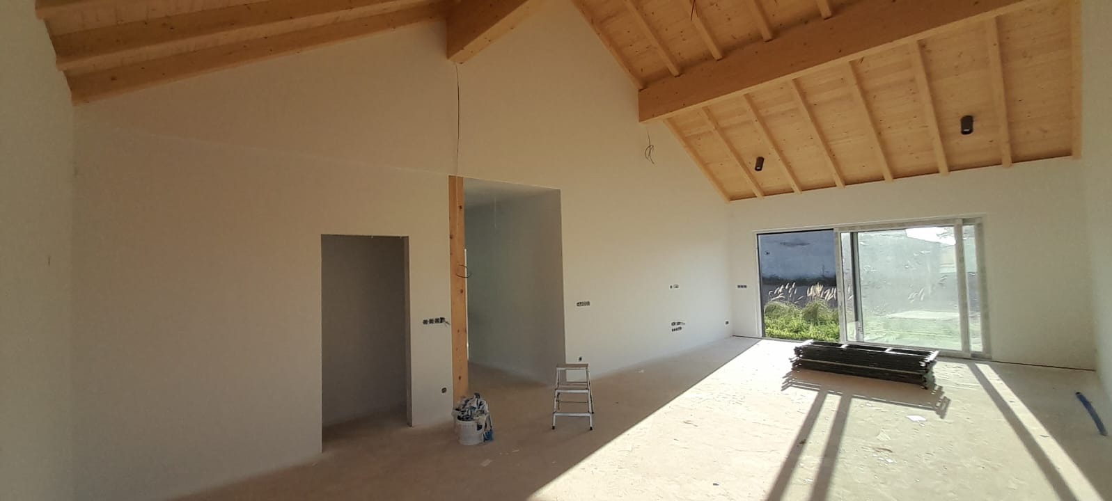 Wnętrze w trakcie prac wykończeniowych: pomalowane ściany, drewniana konstrukcja dachu, surowe otwory drzwiowe, drabina i wiadro z farbą na podłodze.