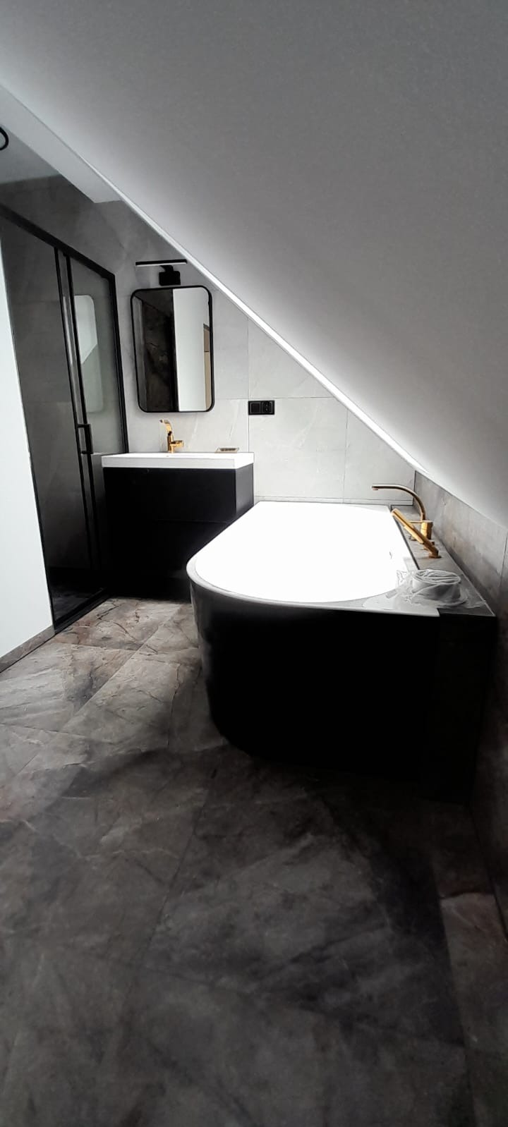 Nowoczesna łazienka na poddaszu z czarną wanną, umywalką i kabiną prysznicową, wykończona szarymi płytkami imitującymi marmur i złotą armaturą.