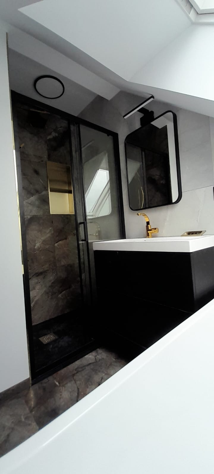 Nowoczesna łazienka na poddaszu z kabiną prysznicową typu walk-in z grafitową armaturą i marmurowym wykończeniem ścian oraz czarną szafką pod umywalką z baterią w kolorze złotym i lustrem w czarnej...