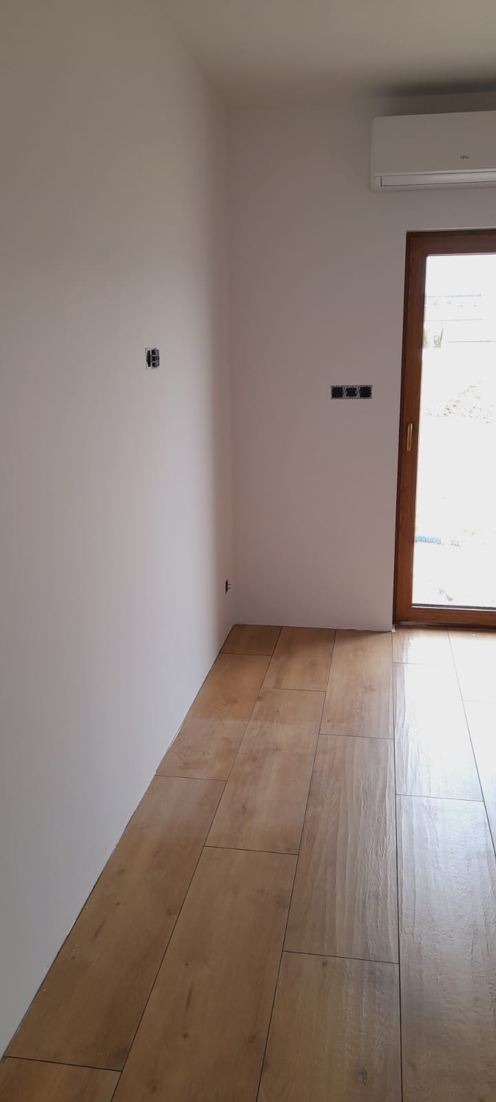 Jasne, drewnopodobne płytki podłogowe ułożone w nowym pomieszczeniu z gładkimi, białymi ścianami, widoczne gniazdka elektryczne i klimatyzator, przez okno widać zieleń.