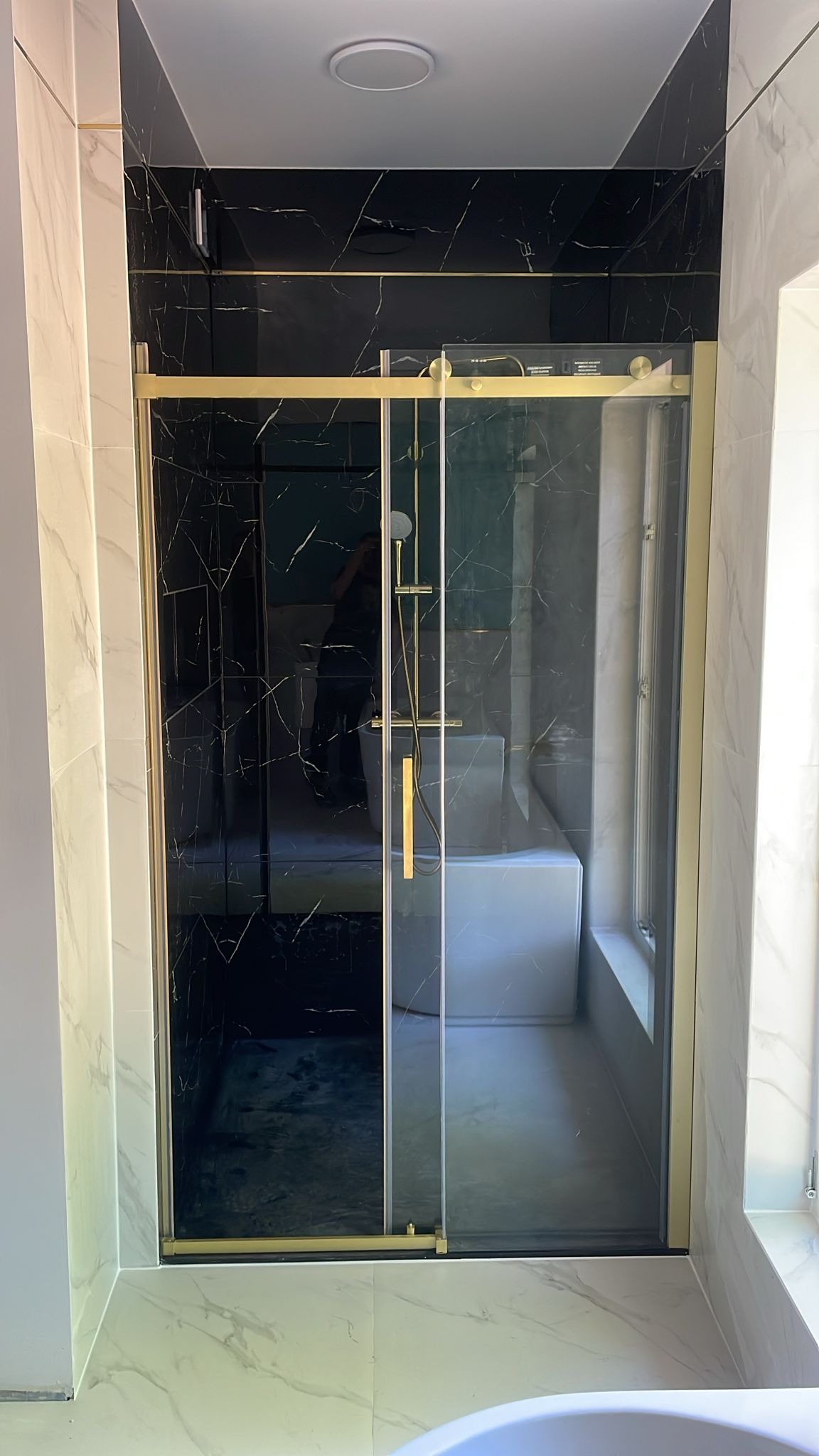 Kabina prysznicowa z przesuwnymi drzwiami, złote profile, czarne marmurowe płytki wewnątrz, białe marmurowe płytki na zewnątrz, widoczna wanna w tle.