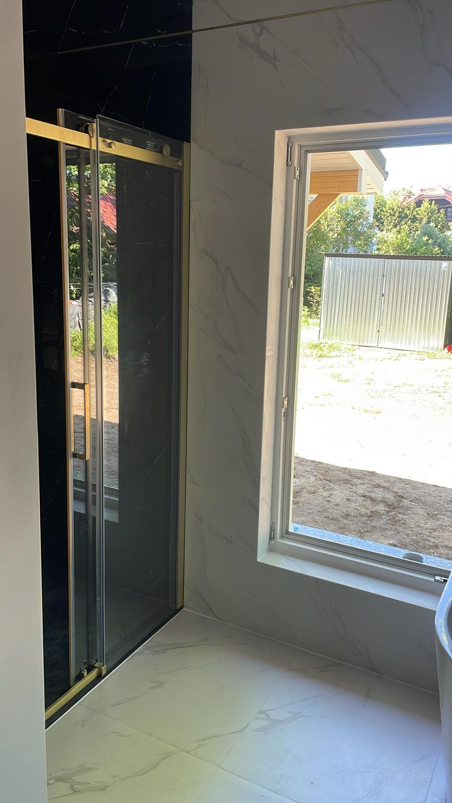 Wykończona łazienka z kabiną prysznicową z czarnym szkłem i złotymi ramami, białe płytki imitujące marmur na ścianach i podłodze, okno z widokiem na ogród.