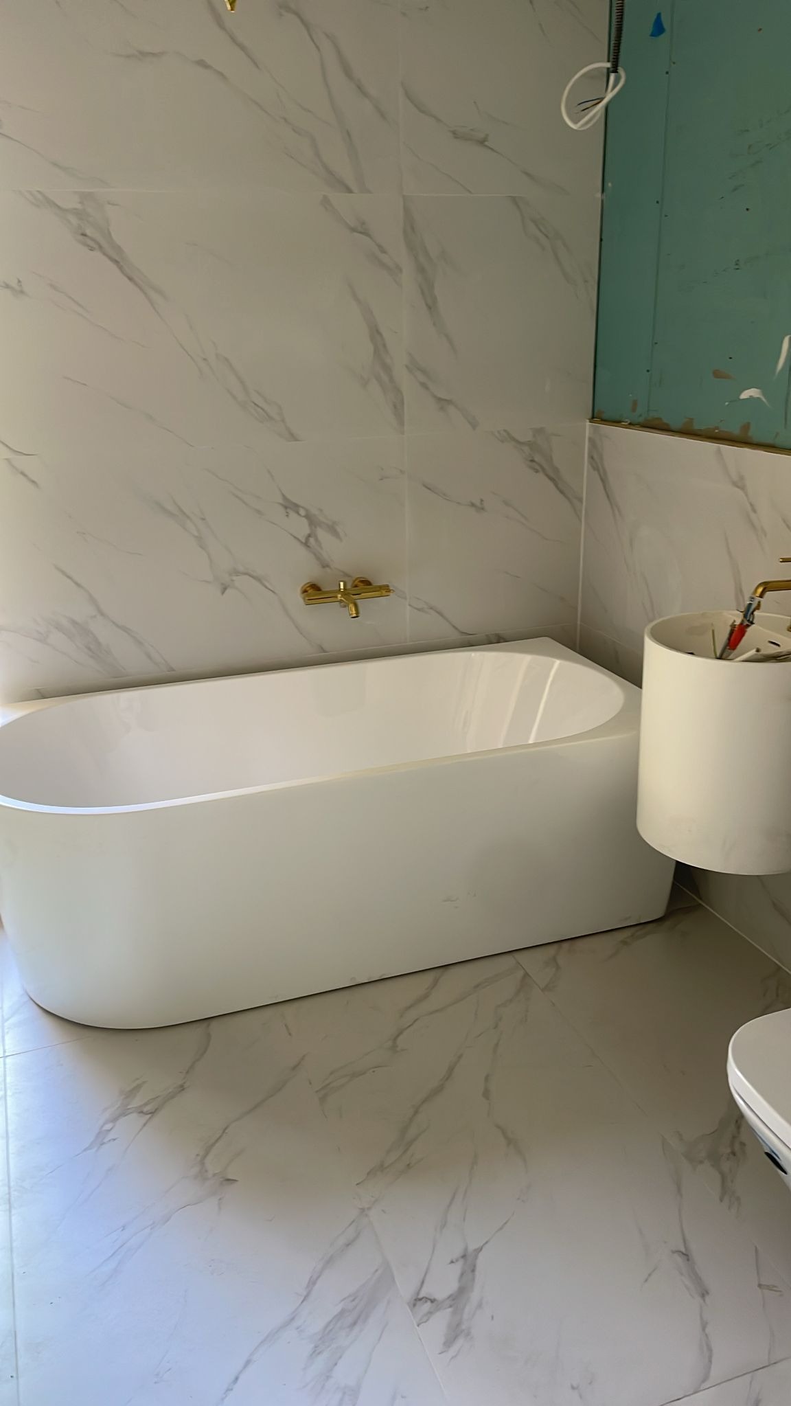 Biała wanna wolnostojąca z baterią w kolorze złota na tle ściany i podłogi wyłożonej płytkami imitującymi marmur. Umywalka w trakcie montażu, widoczne narzędzia i niezabudowana ściana.