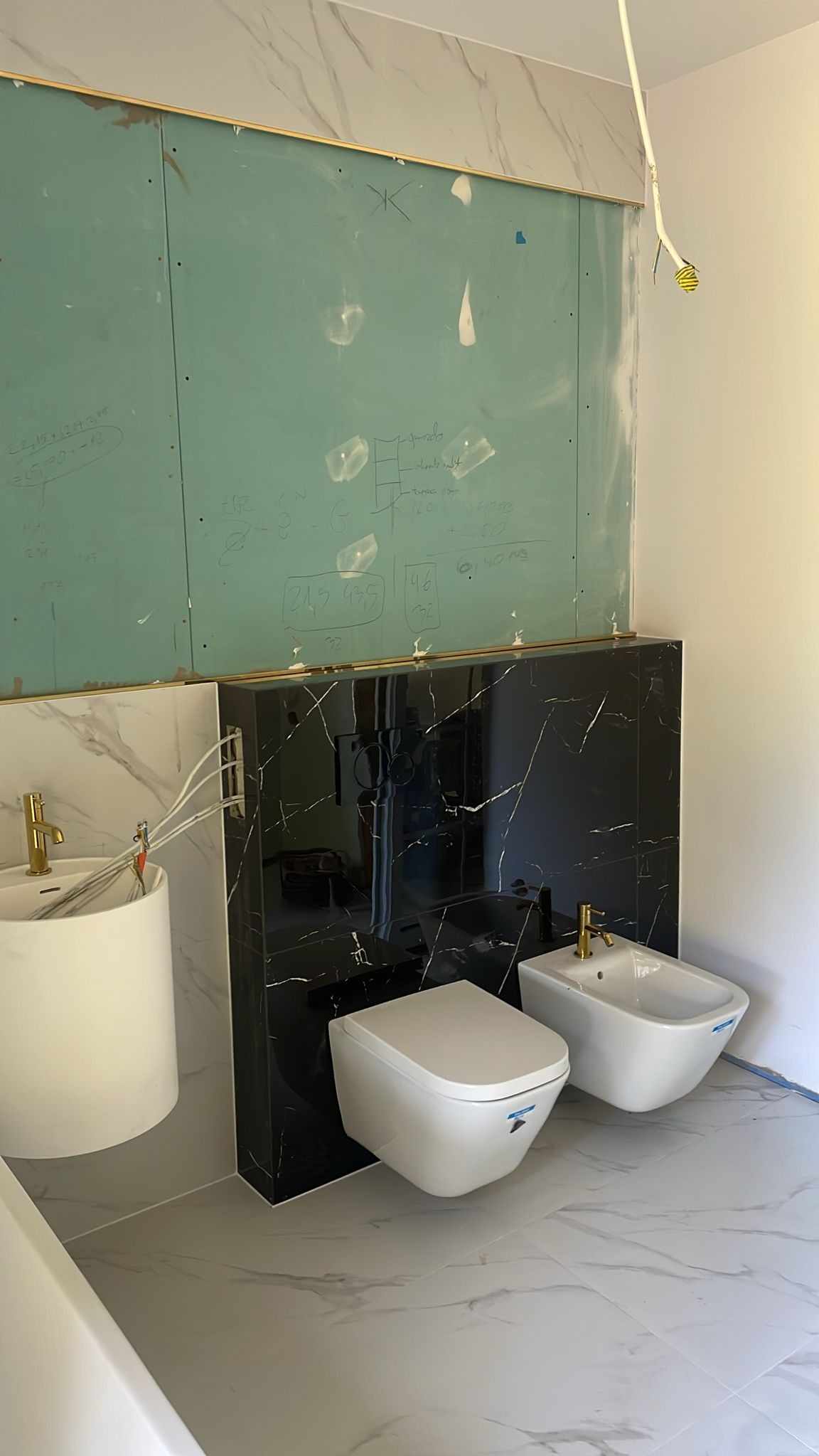 Wnętrze łazienki w trakcie prac wykończeniowych: zamontowana ceramika sanitarna (toaleta i bidet) z armaturą w kolorze złota na tle ściany wyłożonej czarnym marmurem; widoczna także umywalka na tle...