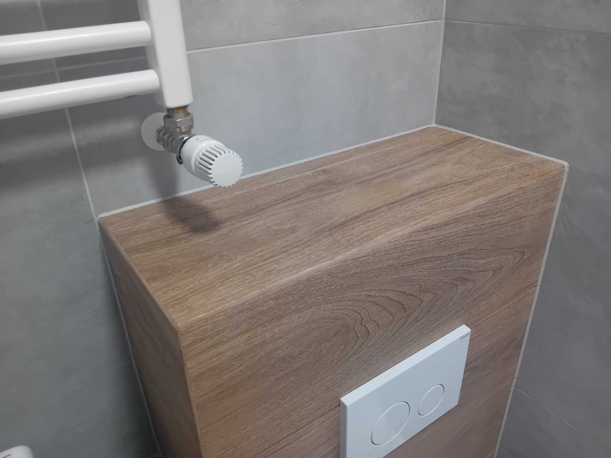 Obudowa stelaża WC z drewnopodobnych paneli, biały grzejnik drabinkowy z termostatem na tle szarych płytek w łazience.