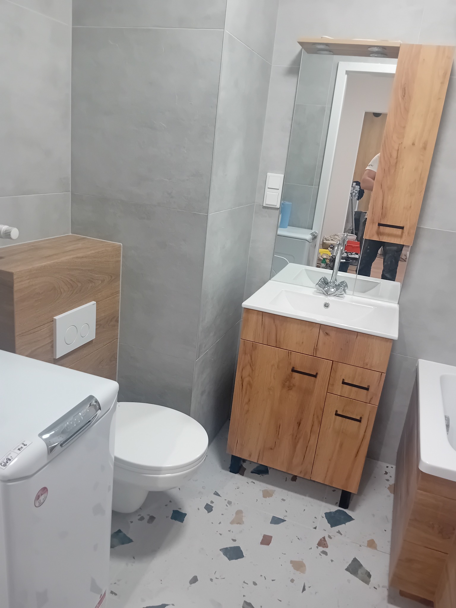 Nowoczesna łazienka z szarymi płytkami, drewnianą szafką pod umywalkę z lustrem, toaletą i pralką, widok na podłogę w stylu lastryko.