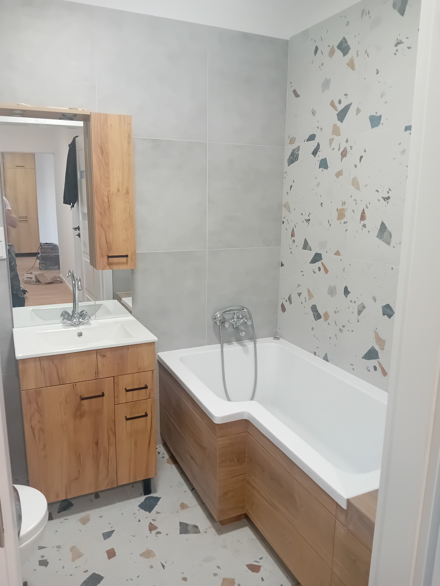 Wykończona łazienka w Bielawie z wanną obudowaną drewnopodobnymi panelami, umywalką z szafką w tym samym stylu, szarą glazurą i ścianą z wzorem terrazzo.