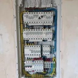 Rozdzielnica elektryczna z zainstalowanymi bezpiecznikami i okablowaniem, widoczne etykiety opisujące obwody, zamontowana w ścianie w trakcie wykończenia.