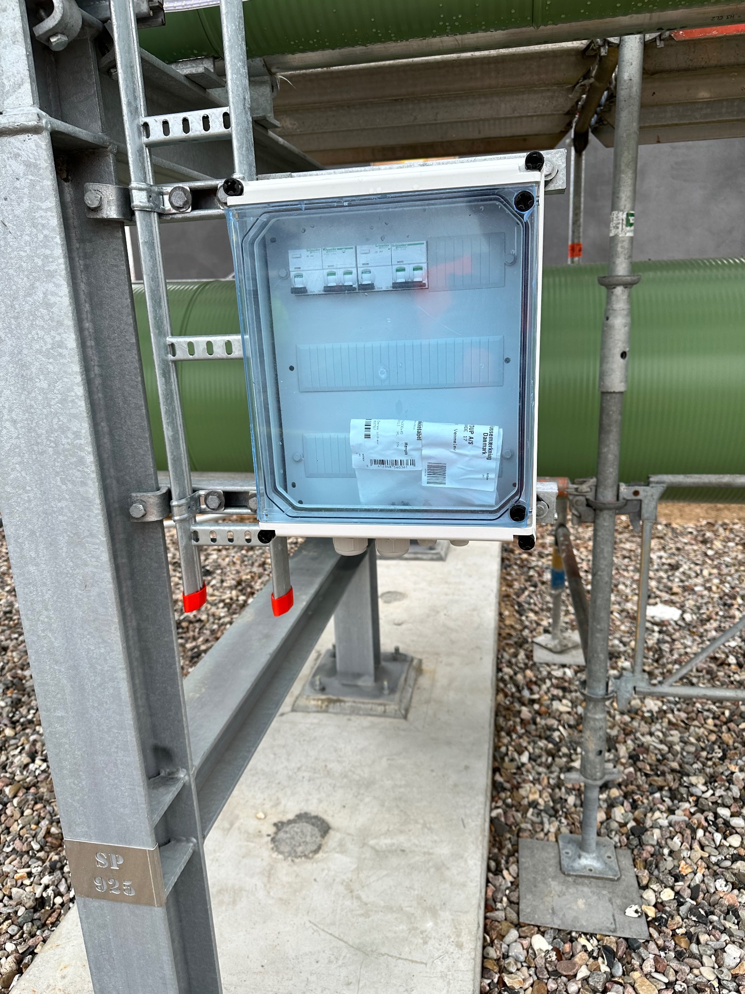 Skrzynka elektryczna z wyłącznikami zamontowana na metalowej konstrukcji wsporczej na zewnątrz budynku, widoczne etykiety i naklejki informacyjne wewnątrz skrzynki.