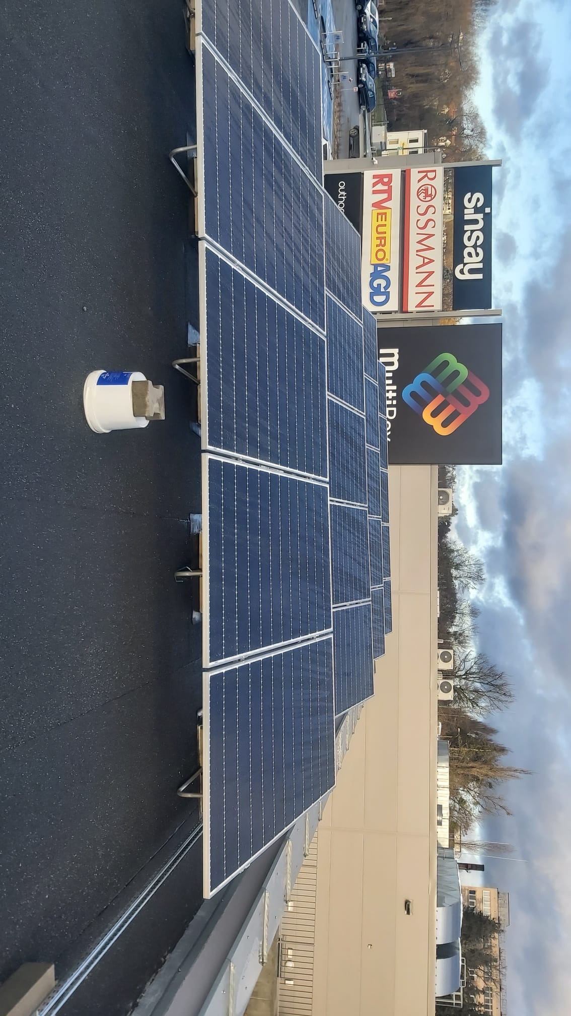 Rząd paneli słonecznych na dachu budynku z widocznymi szyldami sklepów w tle oraz pojemnikiem z betonem obok pierwszego panelu.