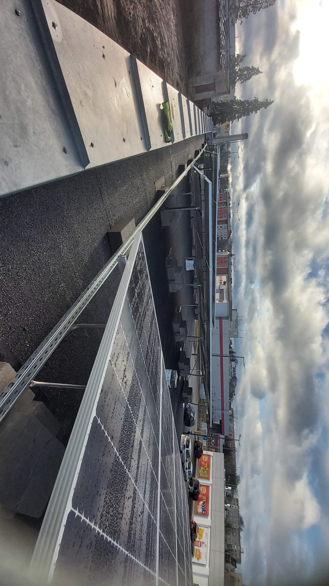 Widok z góry na panele słoneczne z kroplami wody, zamontowane na dachu budynku z parkingiem i sklepem w tle pod zachmurzonym niebem.