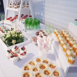 Słodki stół z różnorodnymi deserami: mini bezy z owocami, puddingi w kieliszkach, lizaki bezowe, mini serniczki z karmelem, zielone napoje w stożkowych szklankach, ułożone na białych podestach...