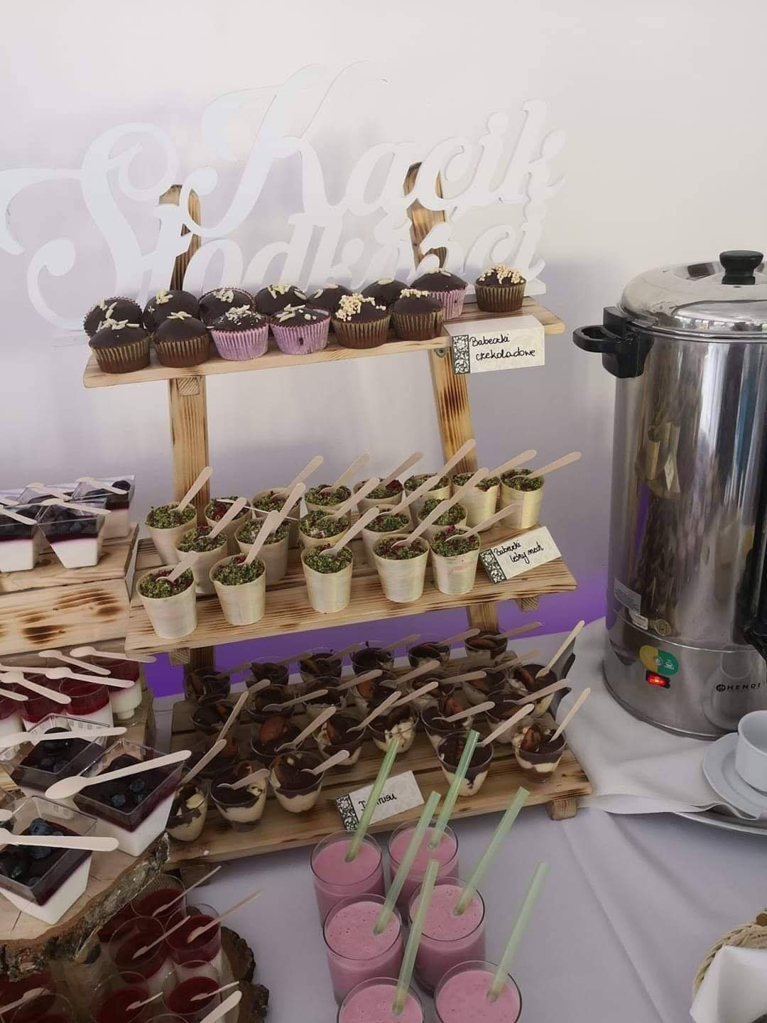 Stół z deserami na drewnianym regale: babeczki czekoladowe, desery w kubeczkach z drewnianymi łyżeczkami, smoothie z zielonymi słomkami i metalowy podgrzewacz do napojów w tle.