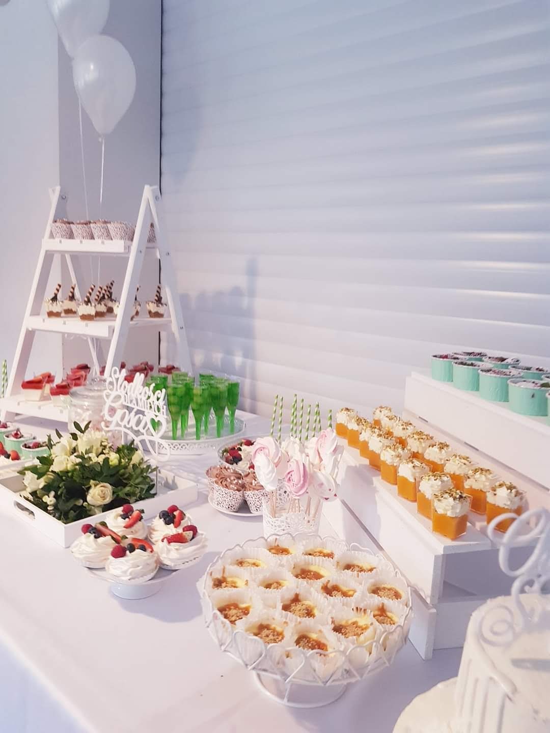 Stół z różnorodnymi deserami: babeczki z owocami, mini serniczki, musy w kieliszkach, lizaki bezowe i zielone napoje, całość w białej kolorystyce z elementami dekoracyjnymi.