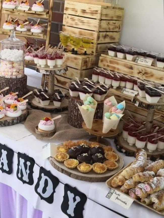 Słodki stół z różnorodnymi deserami, w tym mini tartaletki, panna cotta, bezy i rurki francuskie, zaprezentowany na drewnianych podkładkach i skrzynkach, z dekoracyjnym napisem 'ANDY' w tle.