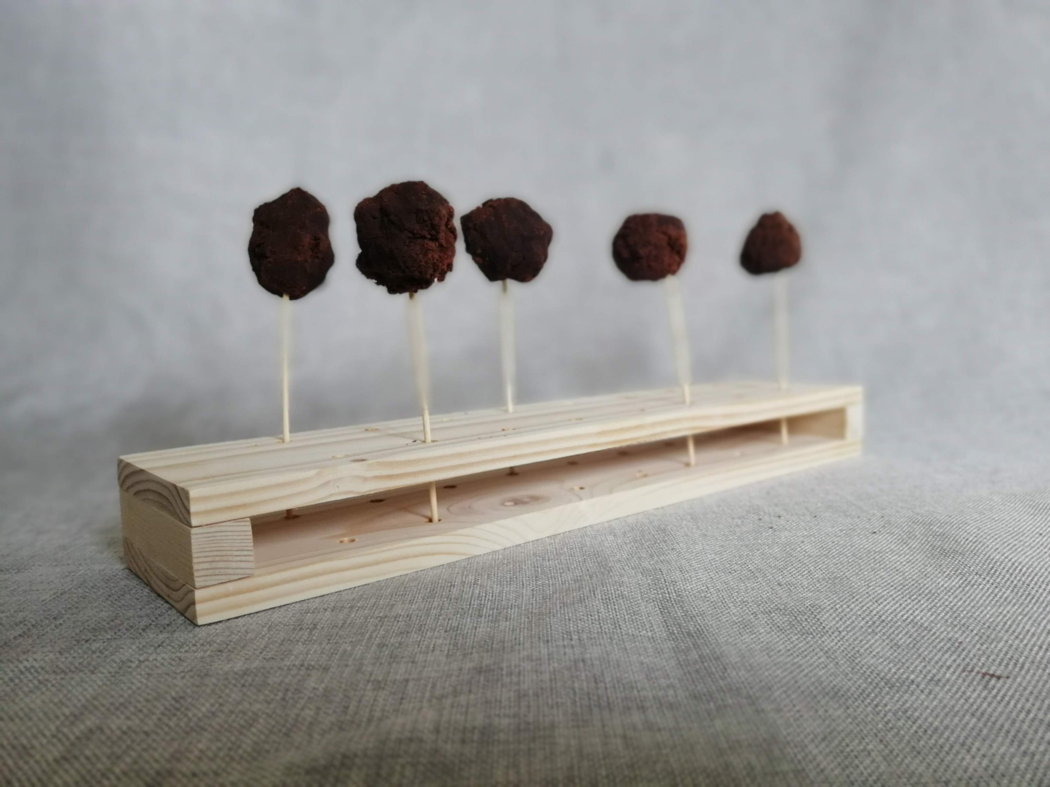 Pięć czekoladowych trufli na patyczkach, umieszczonych w drewnianej podstawce z otworami, na tle szarej tkaniny.