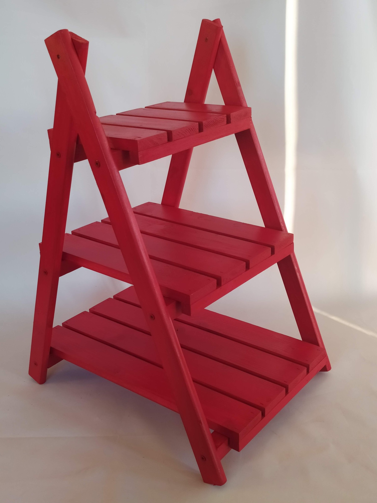 Czerwony, drewniany regał drabinkowy z trzema półkami na białym tle, widoczne ślady pędzla na farbie.