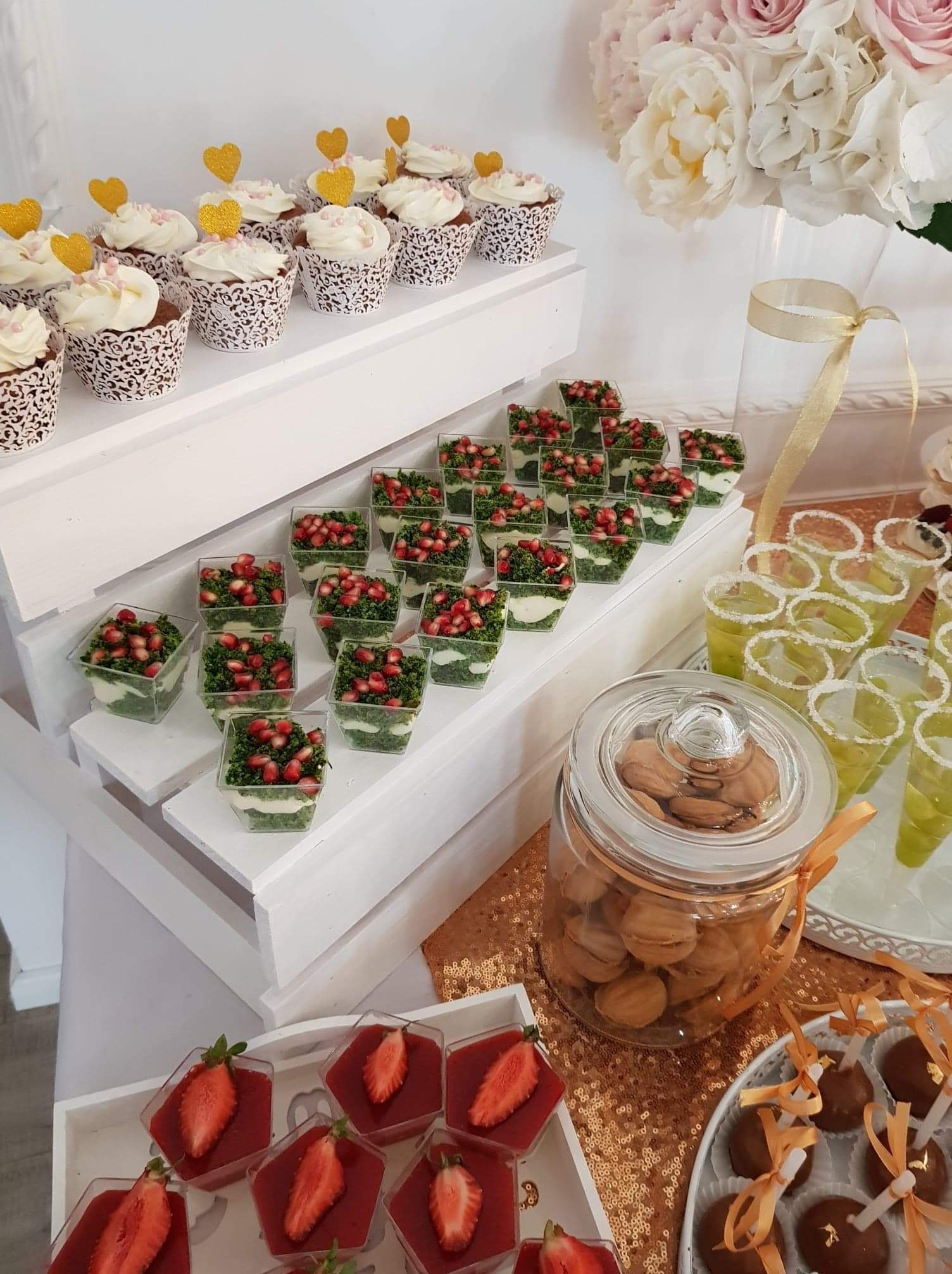 Dekoracyjny stół z różnorodnymi przekąskami i deserami, takimi jak babeczki z serduszkami, sałatki z granatem w małych pojemnikach, galaretki z truskawkami i ciasteczka w słoiku, przygotowany...