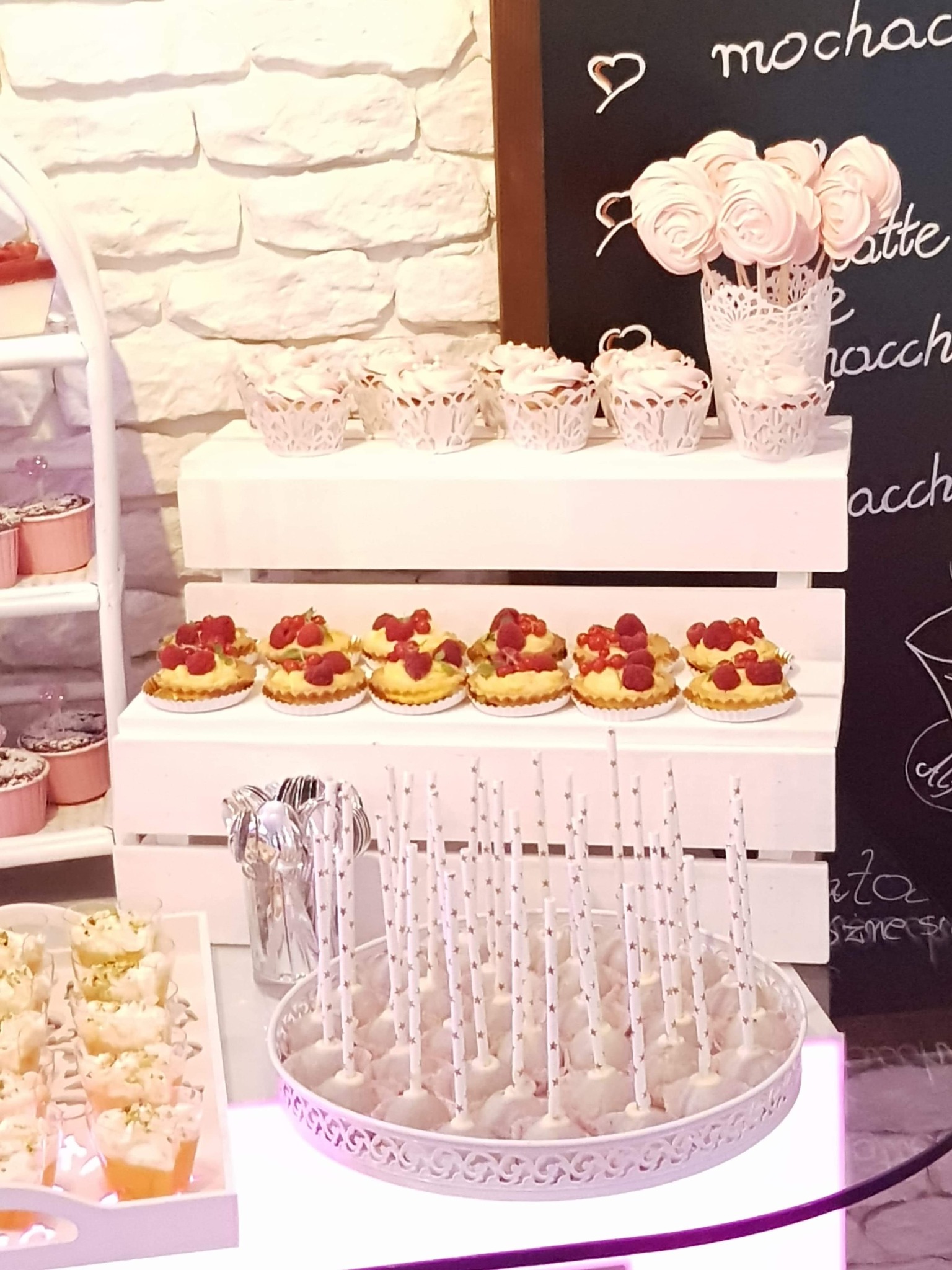 Słodki stół z babeczkami, tartaletkami z owocami i lizakami bezowymi na białych, drewnianych półkach, z tablicą kredową w tle z napisem 'mocha'.