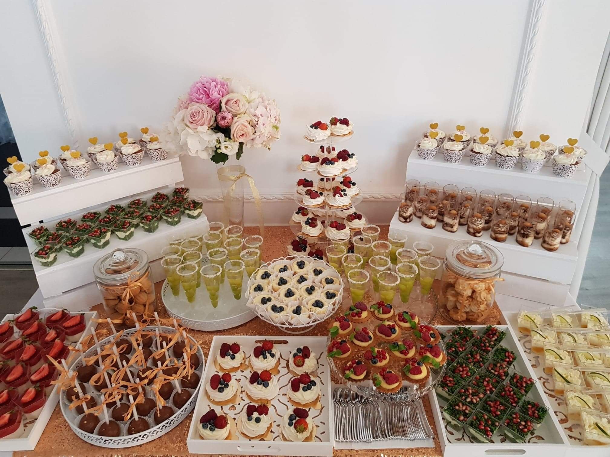 Bogato zastawiony stół z deserami i napojami na przyjęciu, z różnorodnymi słodkościami, od babeczek z serduszkami, przez mini-desery z owocami, po ciasta i poncz w kieliszkach, całość udekorowana...