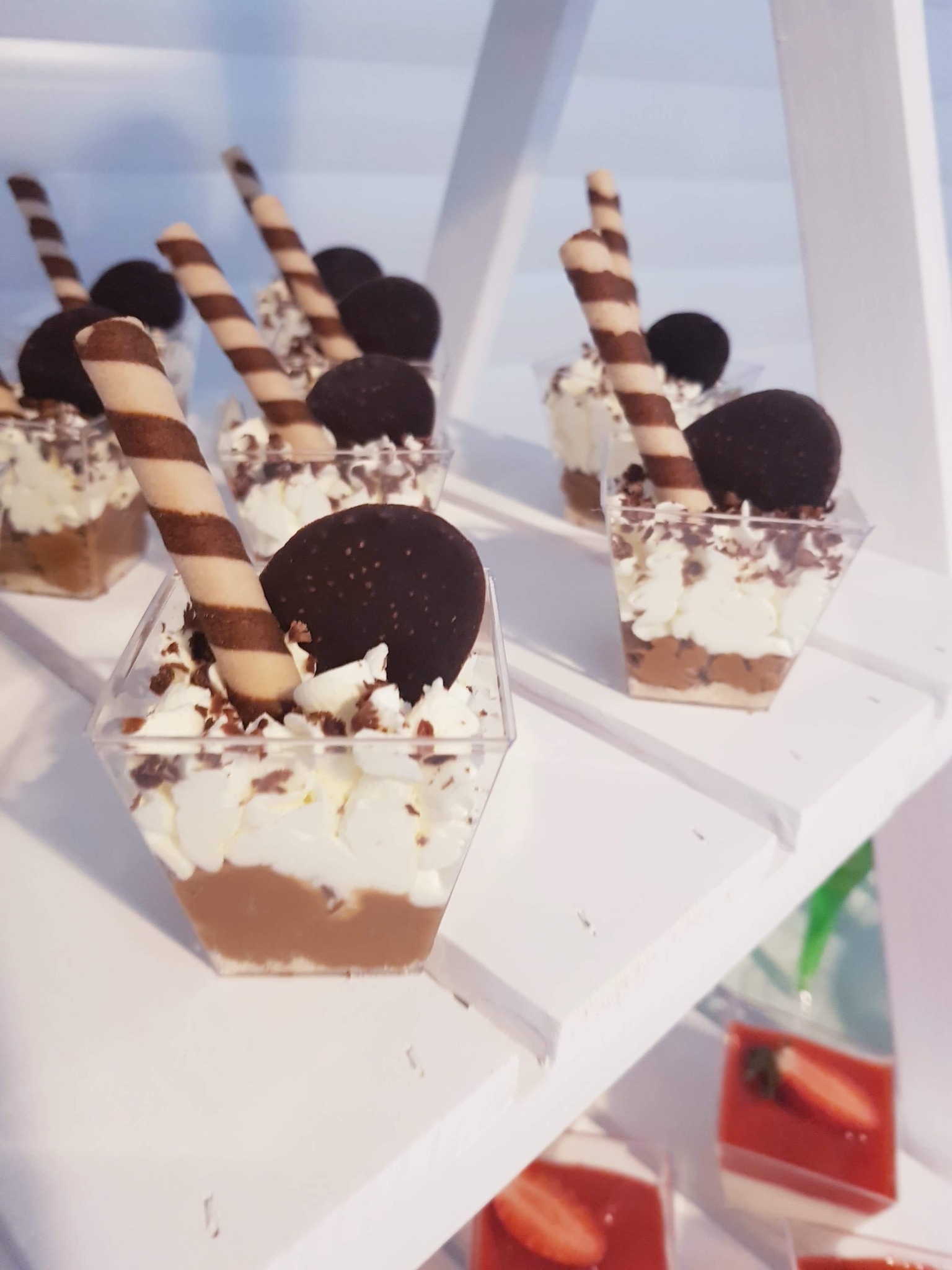 Desery w przezroczystych pojemnikach, ułożone na białym regale, z czekoladowymi ozdobami i rurkami waflowymi.